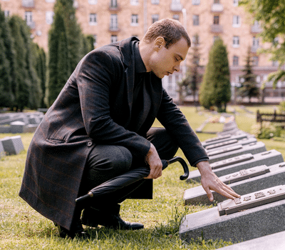 Grieving man in dark funeral attire kneeling beside a grave and touching a memorial plaque during a graveside funeral service