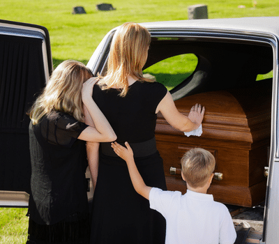 Kids are wearing in funeral attire comforting a grieving woman beside a wooden coffin inside a hearse at an outdoor funeral service